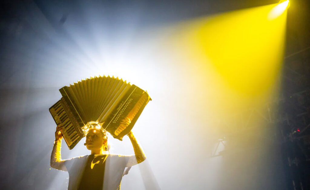 Person on stage playing an accordion, bathed in dramatic yellow stage lighting with a smoky background.