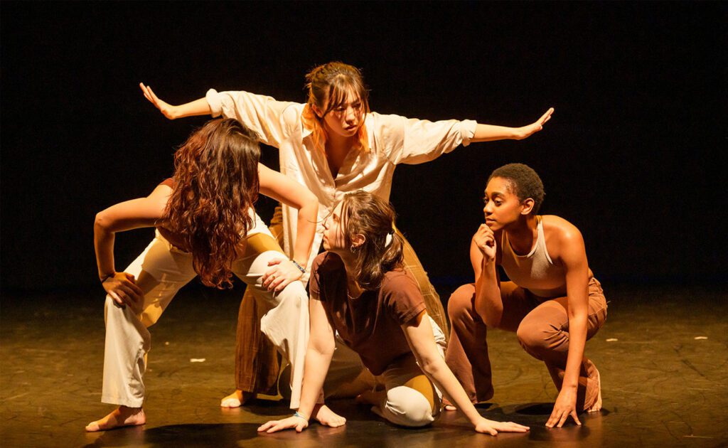 Four dancers in various earth-toned outfits perform an expressive contemporary dance on a dimly lit stage.