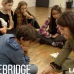 A group of children paying attention to a teacher during a lesson in a classroom setting, with "spacebridge la mama experimental theatre club" signage in the background.