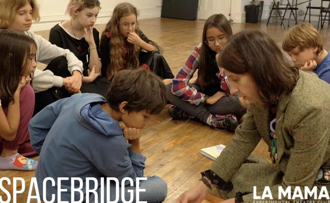 A group of children paying attention to a teacher during a lesson in a classroom setting, with "spacebridge la mama experimental theatre club" signage in the background.