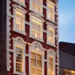A narrow, red brick building with white window frames, illuminated by warm interior lighting, set between two contrasting structures in a city at dusk.