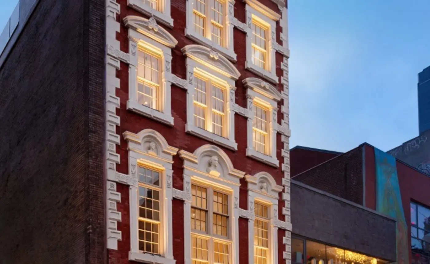 A narrow, red brick building with white window frames, illuminated by warm interior lighting, set between two contrasting structures in a city at dusk.