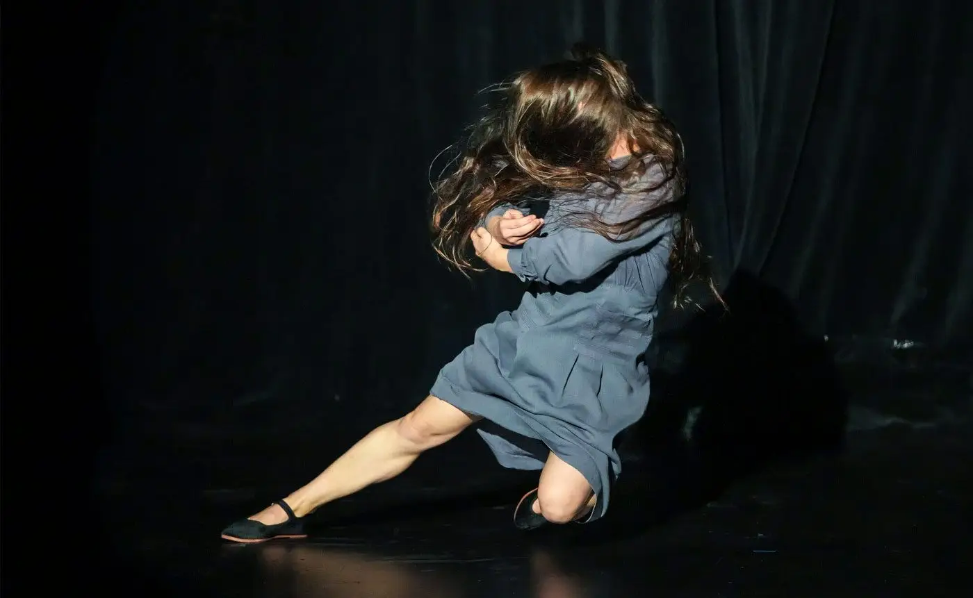 A woman in a blue dress performs a dynamic dance move on stage, her hair flung wildly covering her face.