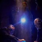 Three men illuminated by a bright light in a dimly lit, brick-walled setting, exuding a mysterious and tense atmosphere.