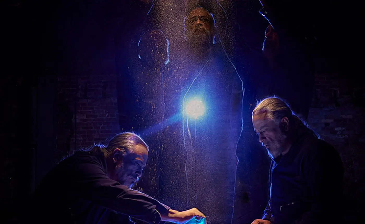 Three men illuminated by a bright light in a dimly lit, brick-walled setting, exuding a mysterious and tense atmosphere.