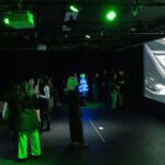 A group of people viewing digital artwork in a dimly lit gallery with green ambient lighting and a large screen displaying abstract visuals.