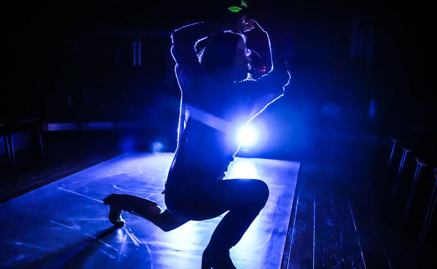 A silhouette of a dancer on stage, highlighted by a bright blue backlight, capturing a dynamic pose with one knee bent and hands above the head.