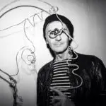 A man in a leather jacket and beanie poses next to a wire art piece of a face, wearing a quirky monocle, in a black and white photograph.