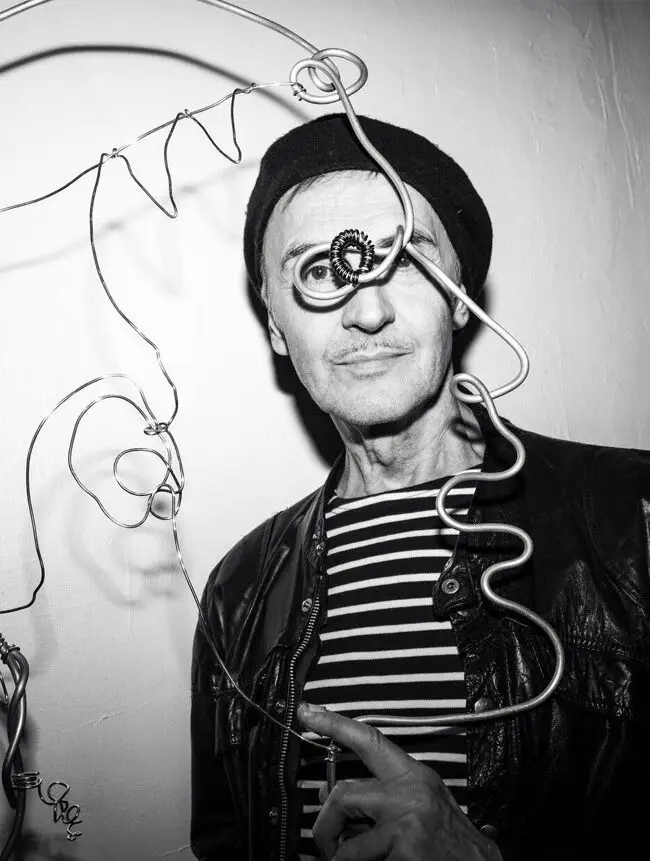 A man in a leather jacket and beanie poses next to a wire art piece of a face, wearing a quirky monocle, in a black and white photograph.