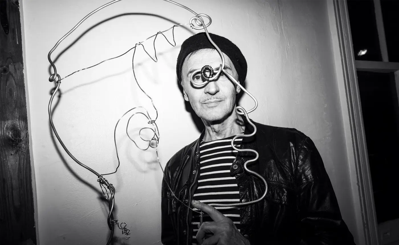 A man in a leather jacket and beanie poses next to a wire art piece of a face, wearing a quirky monocle, in a black and white photograph.