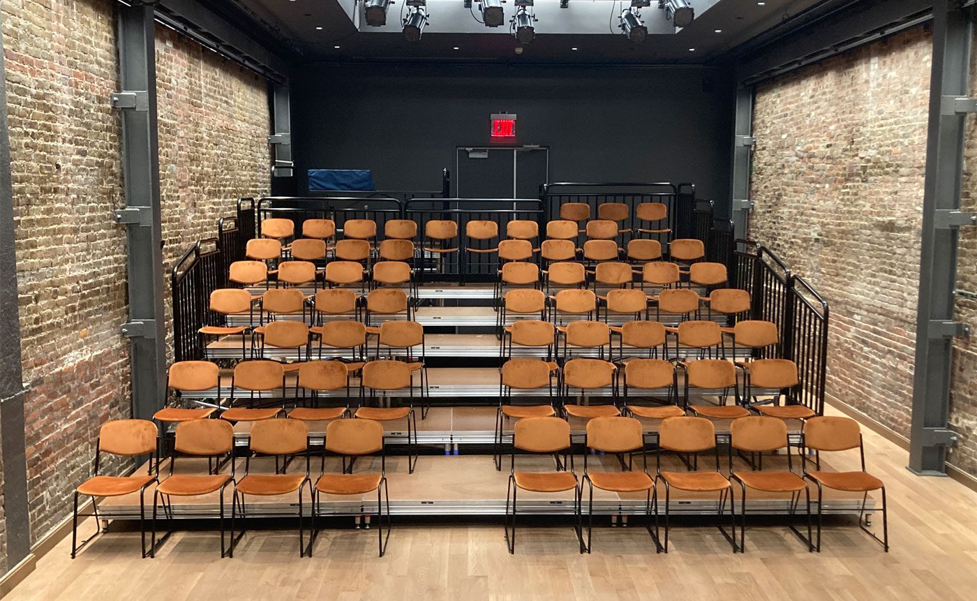 Small, empty theater with rows of orange chairs facing a black stage, flanked by exposed brick walls and dark curtains.