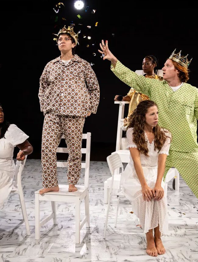 Five actors in eclectic costumes perform on a stage with a white floor, using chairs as props in a dramatic scene.