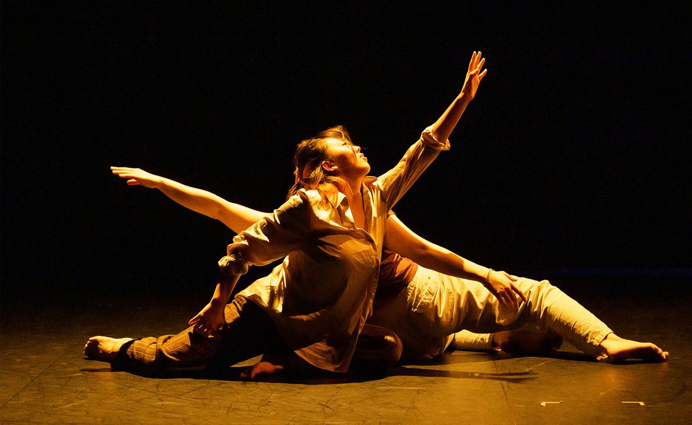 Two dancers in a dramatic pose on a dimly lit stage, one is arching back while the other stretches out an arm, both are wearing flowing costumes.