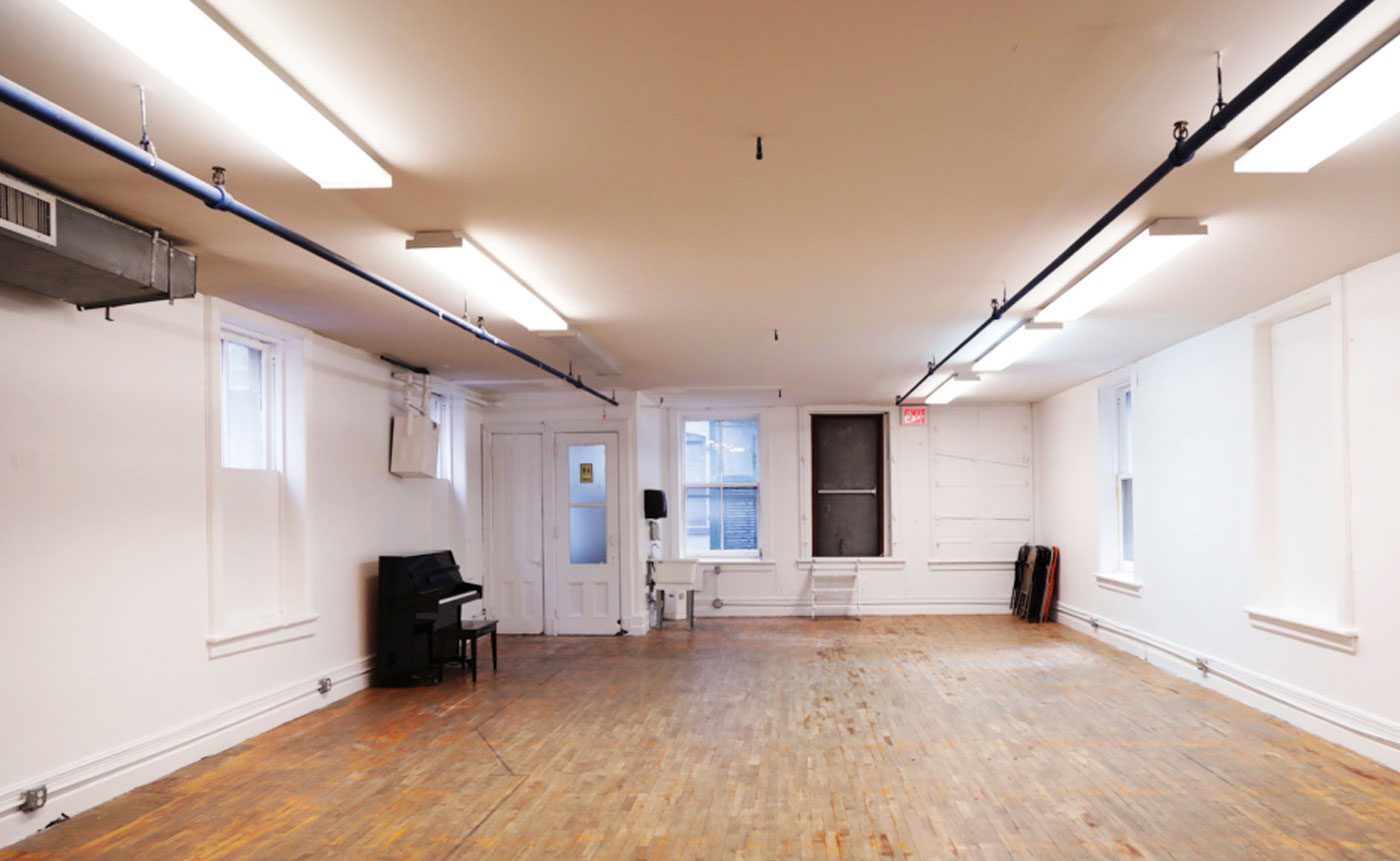 Empty dance studio with wooden floor, white walls, and a black piano in the corner.