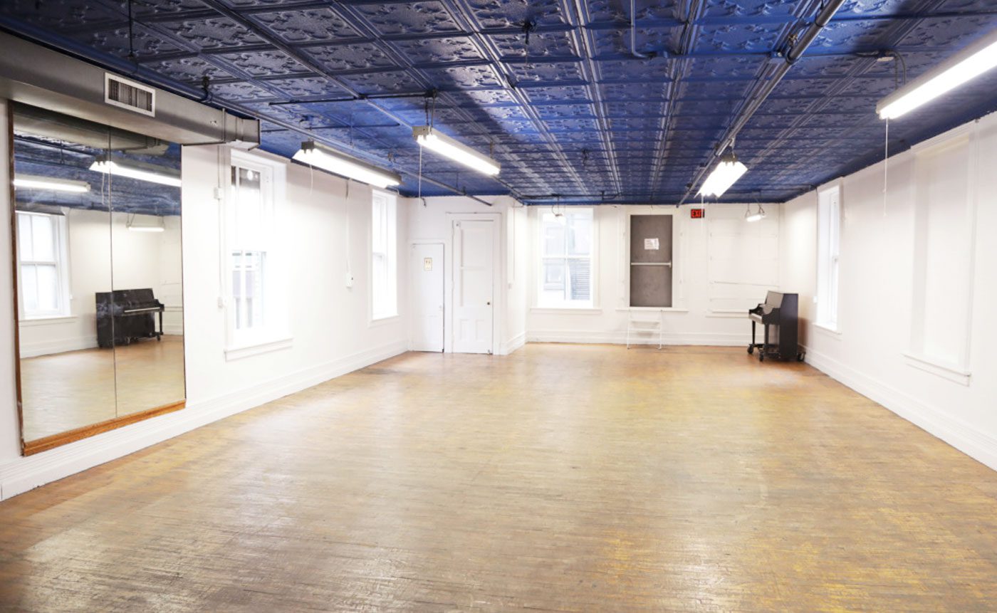 Empty dance studio with wooden floors, mirrored wall on the left, white walls, and a blue decorative ceiling. there is a piano in the corner.