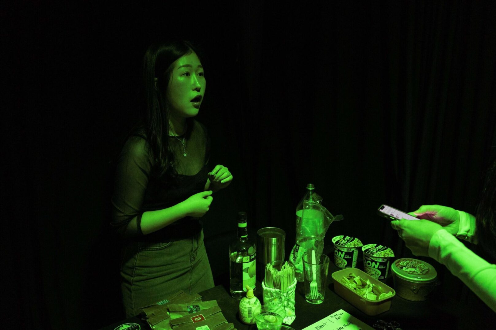 A woman stands in a dimly lit room with a green light, looking surprised as she talks to someone off-camera, surrounded by bottles and various items on a table.