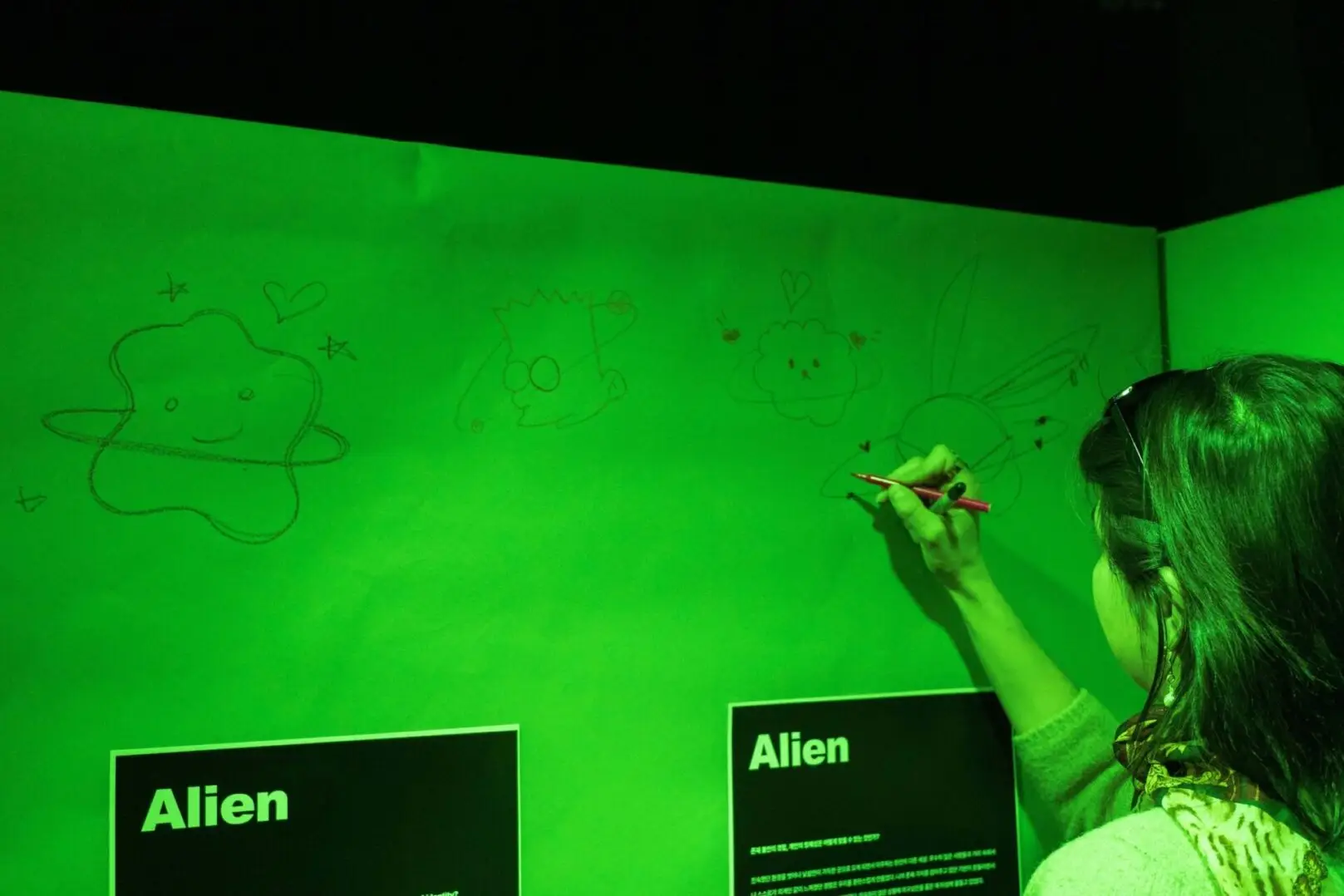 A woman sketches whimsical alien drawings on a green glowing wall illuminated by black light.