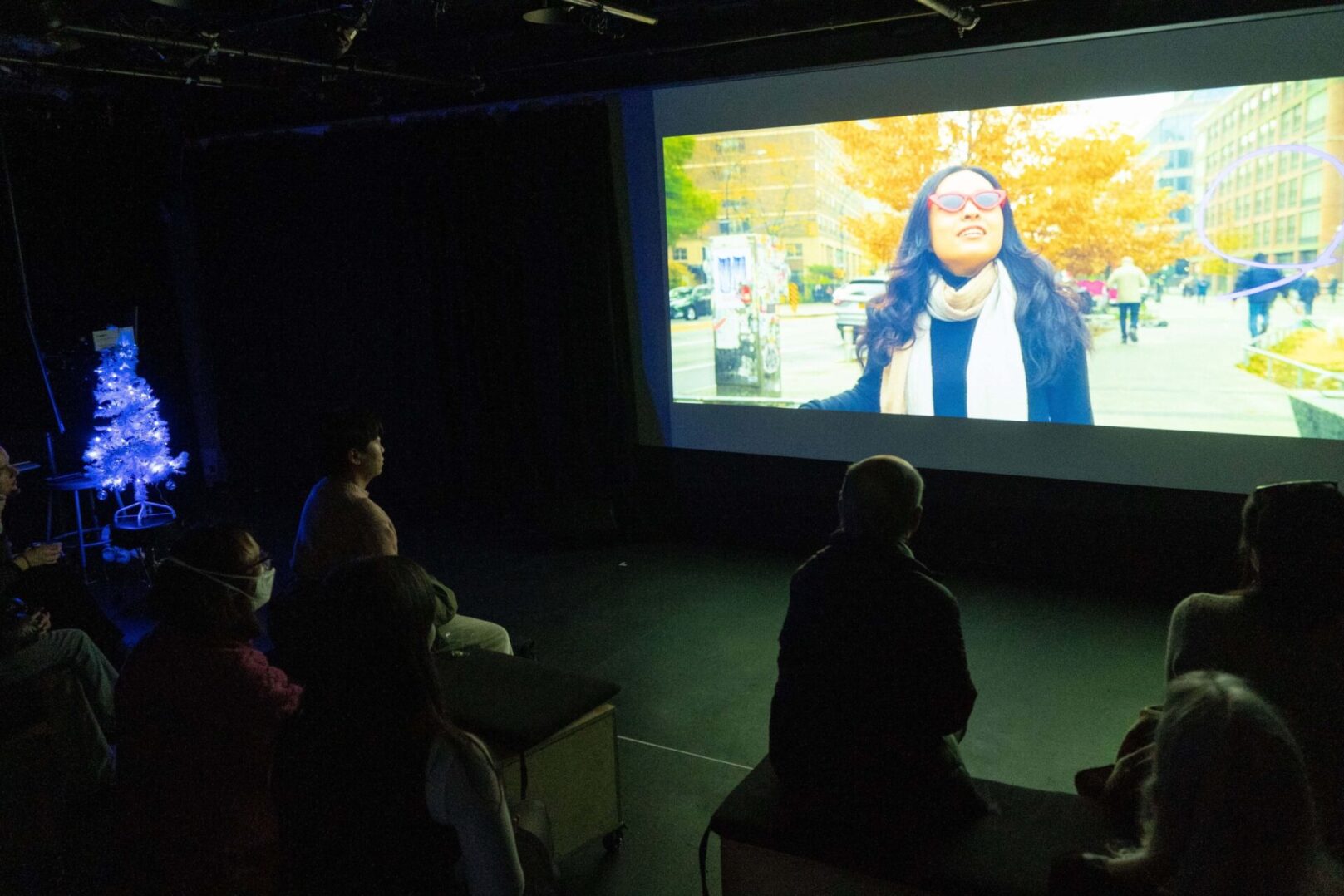 Audience members watching a projection of a woman walking in a city setting on a large screen in a dark room.