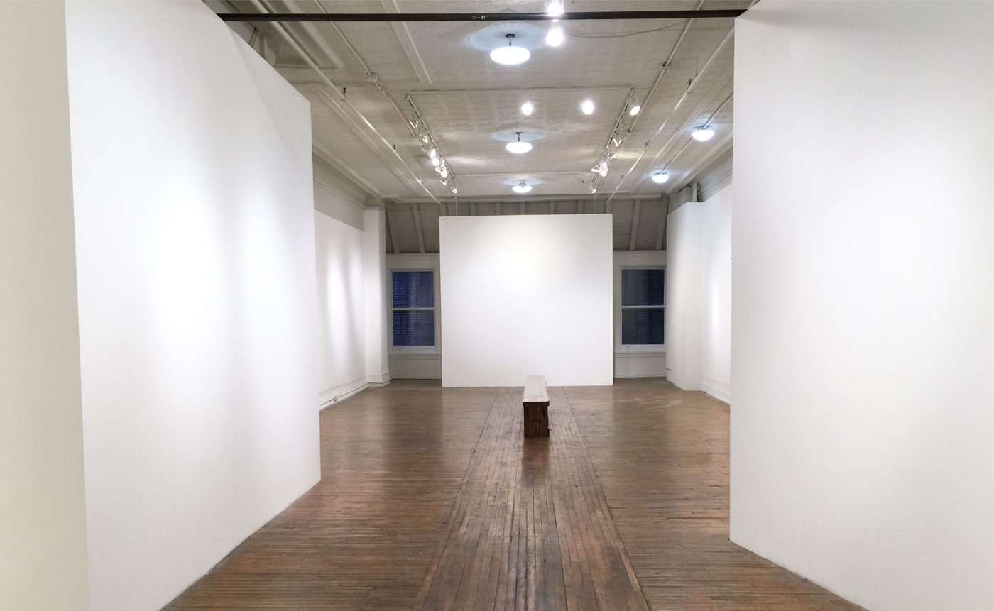 Empty art gallery with white walls, spotlights, a wooden bench in the center, and a large opaque panel at one end.