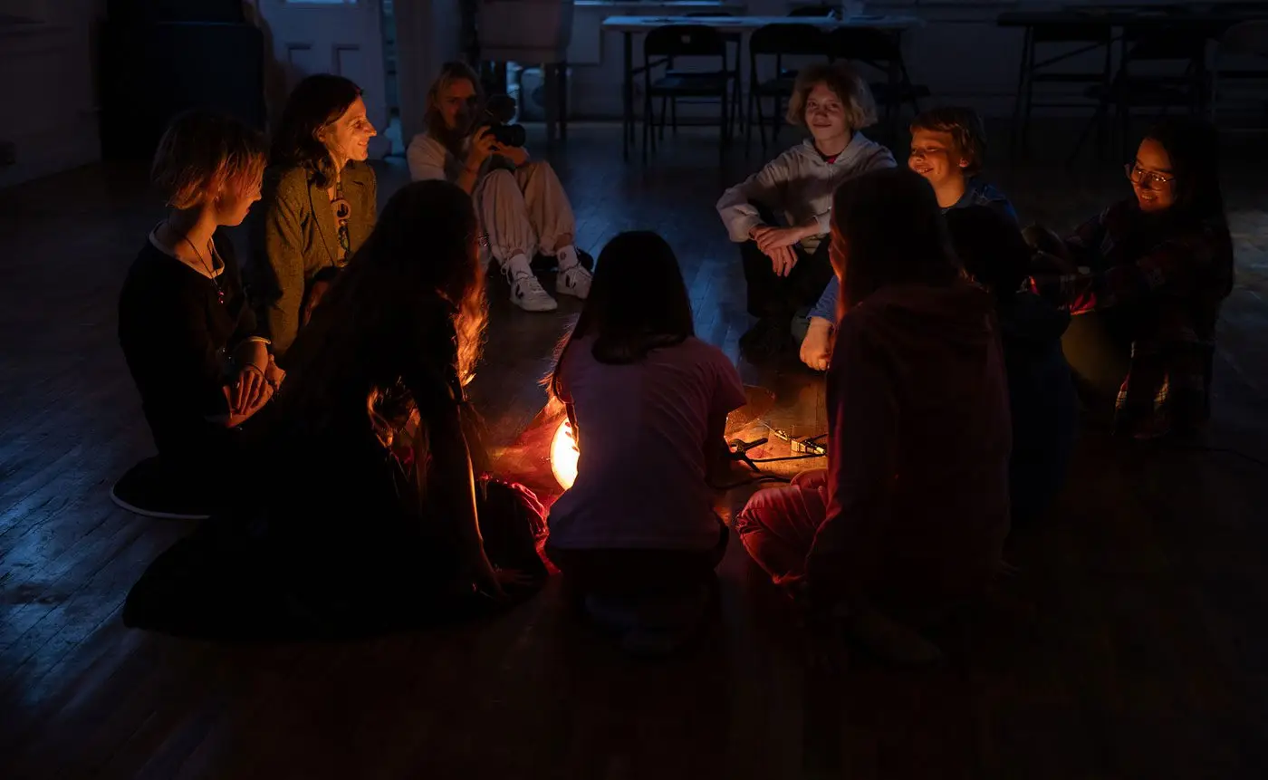 A group of young people sitting in a circle in a dimly lit room, engaging in a conversation around a small artificial campfire.