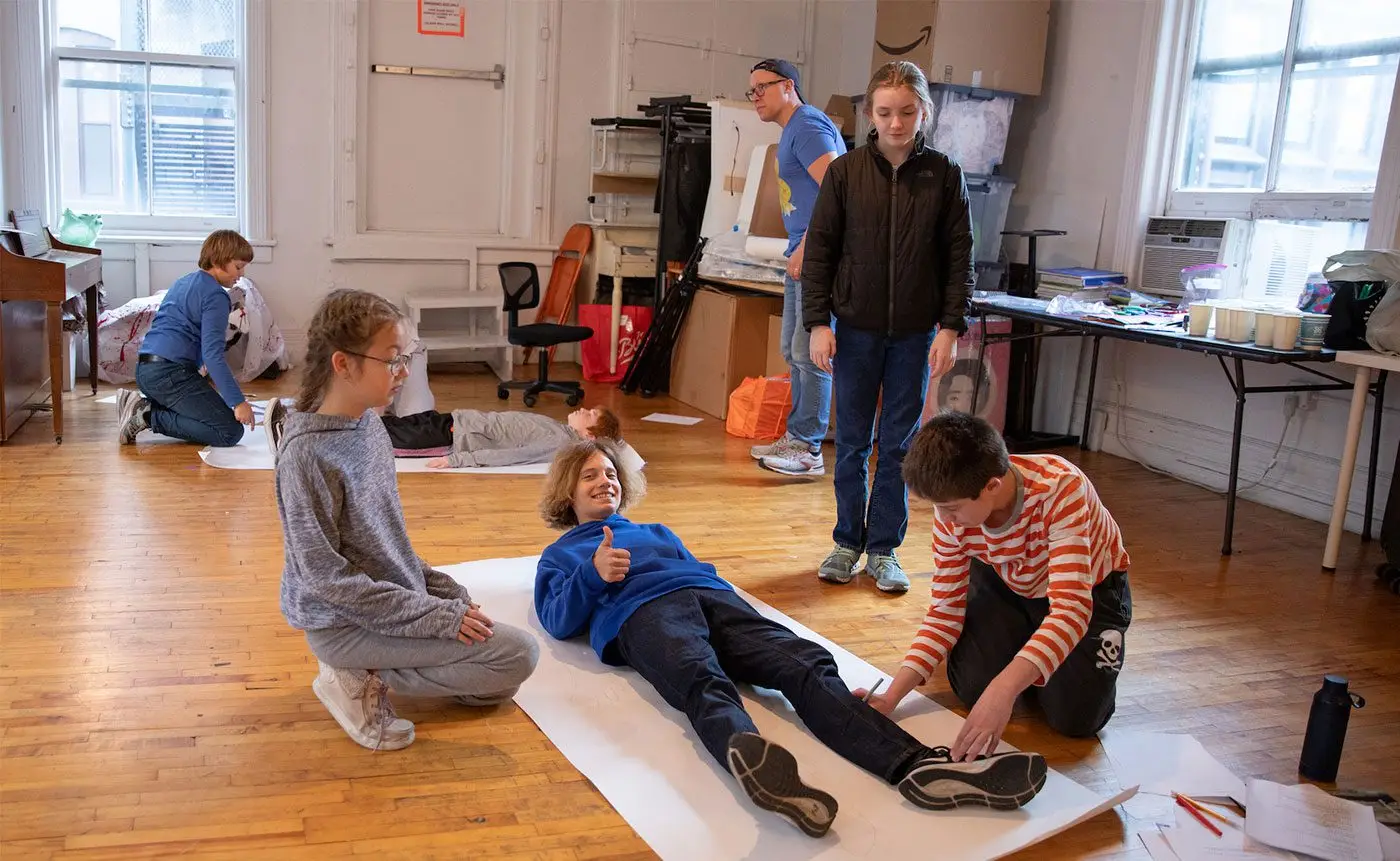 Children participate in an art class, tracing a boy's silhouette on a large sheet of paper in a classroom with other activities in the background.