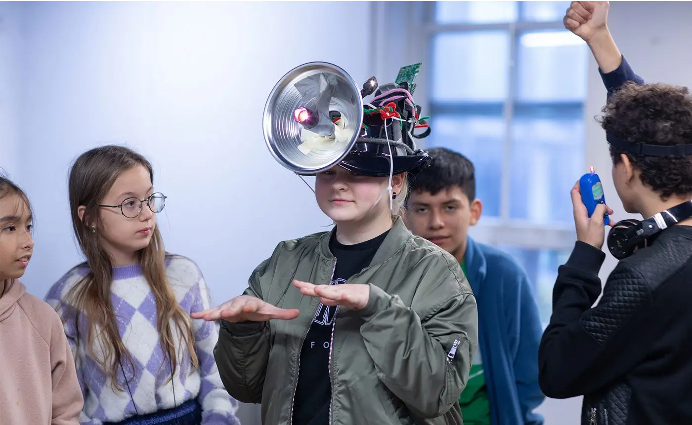 Student wearing a creative helmet adorned with electronic components, demonstrating it to classmates in a classroom setting.