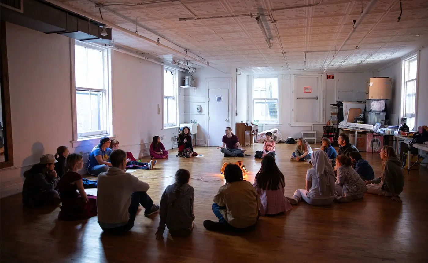 Adults sitting in a circle on a wooden floor in a spacious room, engaged in a group discussion.