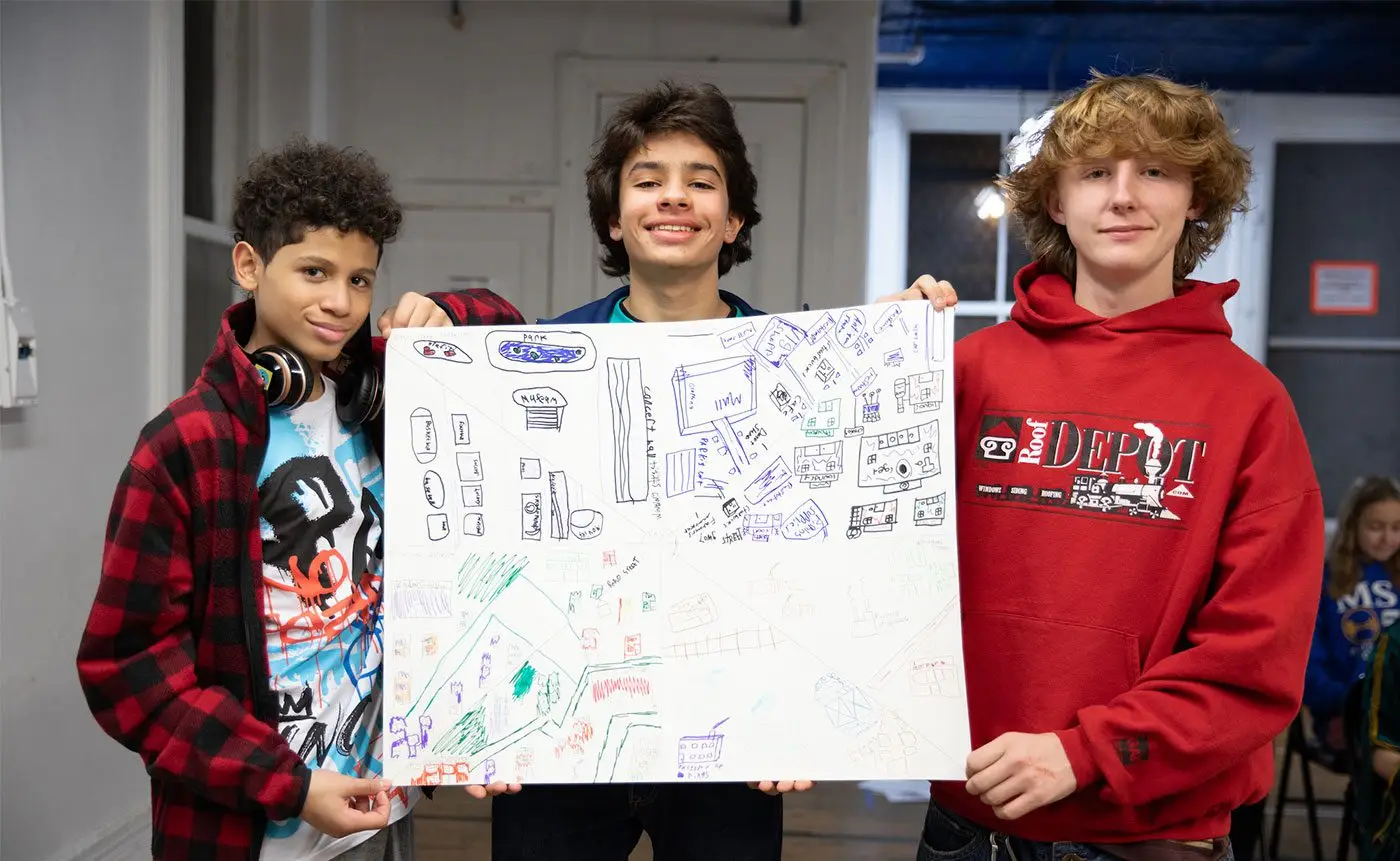 Three boys smiling and holding up a large poster with colorful drawings and text, standing in a room with other people in the background.