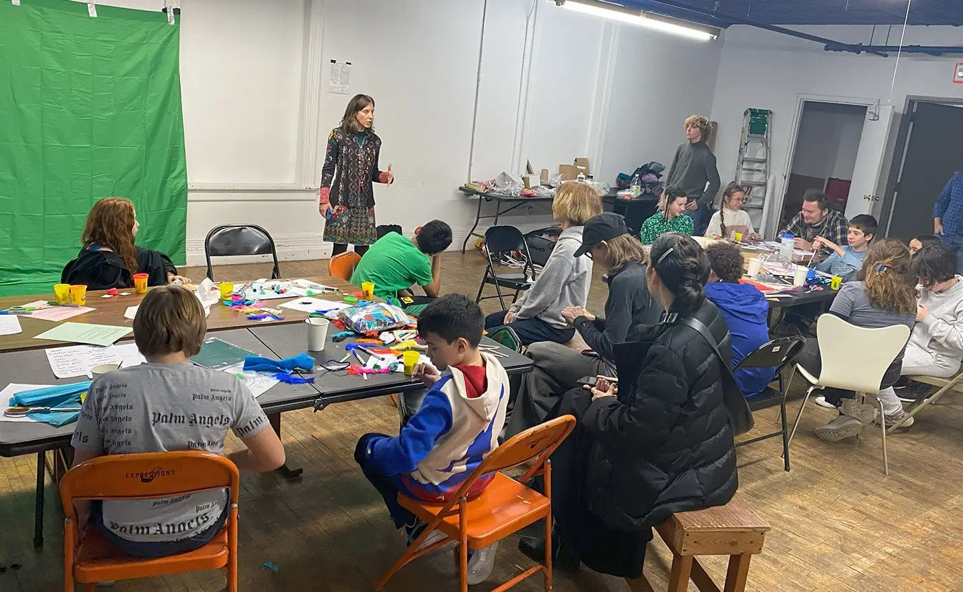 A woman stands teaching a group of children and teens engaged in arts and crafts activities at tables in a room with a green screen.