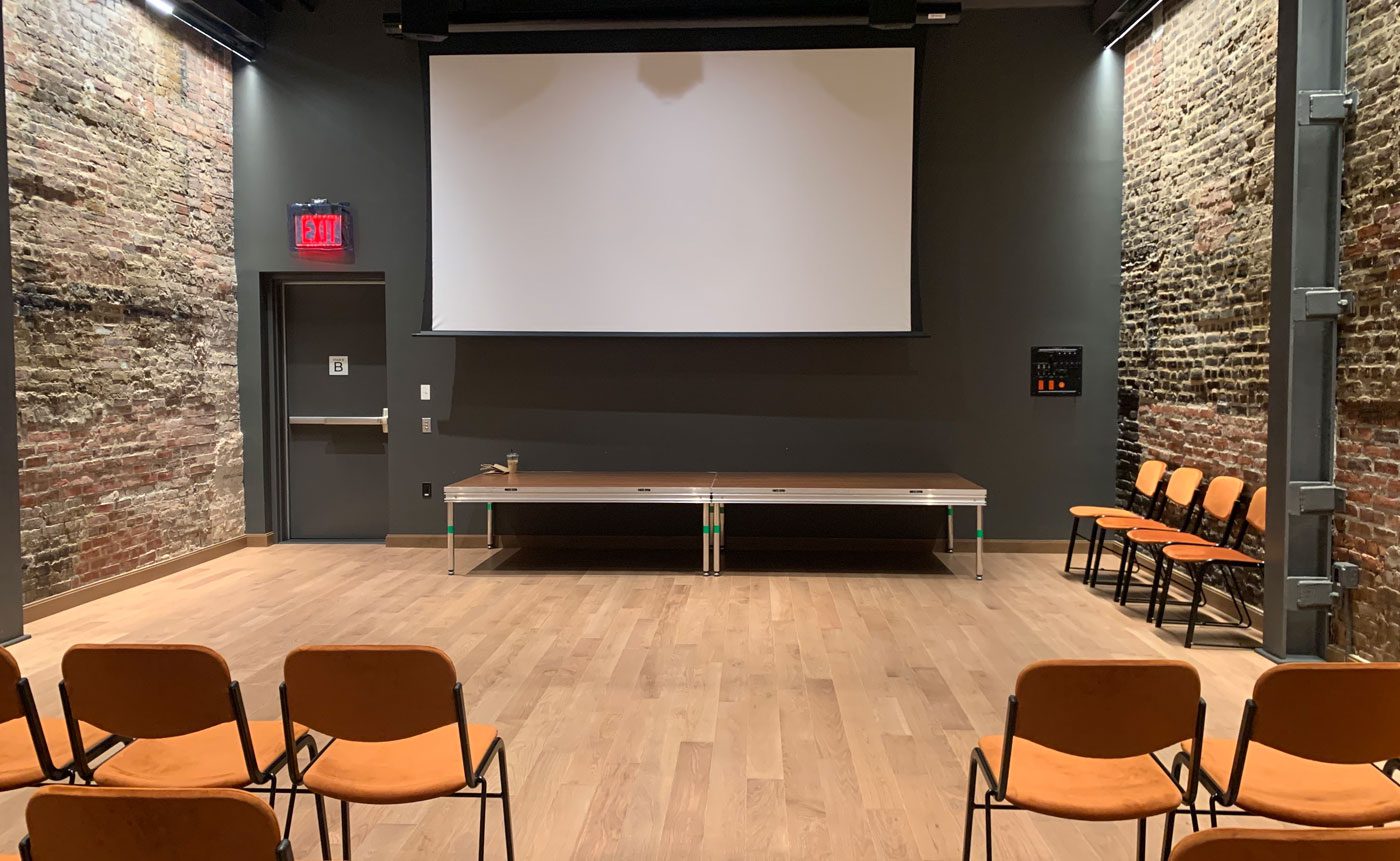 Interior of a modern meeting room with brick walls, featuring a ping pong table, a projector screen, and rows of chairs on wooden flooring.